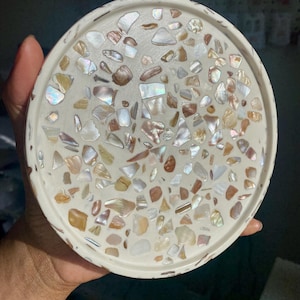 May include: A circular, off-white decorative tray featuring a mosaic pattern. The mosaic is composed of irregularly shaped pieces of iridescent shell in white, tan, and brown hues. The tray is held by a hand.