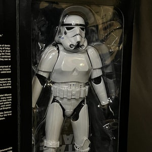 Puede incluir: Una figura de acción de plástico blanco de un Stormtrooper de Star Wars. La figura está en posición de pie con los brazos a los lados. La figura lleva un casco blanco con una visera negra. La figura está empaquetada en una caja de plástico transparente con un fondo negro.