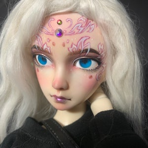 May include: A white haired doll with blue eyes and pink and white face paint. The doll is wearing a black shirt and has a black strap across its chest.