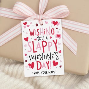 Peut inclure: Un cadeau emballé dans du papier brun clair, orné d'un ruban rayé rose et blanc. Une étiquette cadeau blanche porte l'inscription "Wishing You a Slappy Valentine's Day!" avec des cœurs et des étoiles.