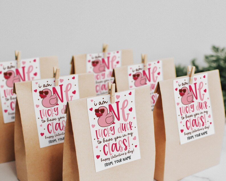 Printable lucky duck Valentine gift tag clipped to a brown paper treat bag, with a pink duck wearing heart sunglasses, bold “ONE LUCKY DUCK” lettering, scattered pink hearts, and gold dots, ideal for teachers to print, cut, and attach to classroom student Valentine favors.