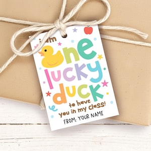 May include: A rectangular white gift tag featuring a yellow rubber duck illustration and colorful text that reads: I am one lucky duck to have you in my class! FROM: YOUR NAME. The tag is decorated with small stars, dots, and a red apple.