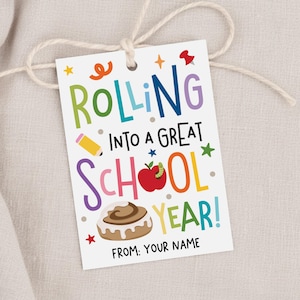 Może przedstawiać: Prostokątna biała zawieszka prezentowa z otworem i białym sznurkiem, zawierająca kolorowy napis "ROLLING INTO A GREAT SCHOOL YEAR!" oraz ilustracje żółtego ołówka, czerwonego jabłka z zielonym robakiem i brązowej bułeczki cynamonowej.