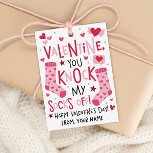 May include: A Valentine's Day gift tag with the text "VALENTINE, YOU KNOCK MY SOCKS OFF!" in red and pink. The tag features illustrations of socks and hearts. The tag is tied to a wrapped gift with pink twine.