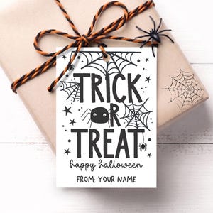 Puede incluir: Una etiqueta de regalo rectangular blanca con texto negro que dice TRICK OR TREAT, happy halloween y FROM: YOUR NAME. El diseño incluye ilustraciones de telarañas negras, estrellas pequeñas y una araña dibujada con dos ojos.