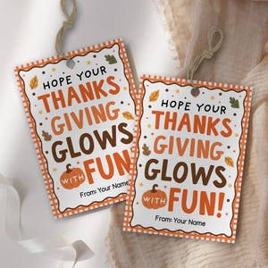 Puede incluir: Dos etiquetas de regalo rectangulares de papel con un centro blanco y un borde a cuadros naranja y blanco. El texto dice HOPE YOUR THANKSGIVING GLOWS WITH FUN! y From: Your Name. Cada etiqueta presenta una pequeña calabaza naranja.