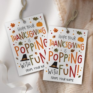 Könnte beinhalten: Zwei weiße Thanksgiving-Geschenkanhänger mit dem Text "Hope Your Thanksgiving is Popping with Fun!" in bunten Buchstaben. Die Anhänger zeigen herbstliche Illustrationen und sind mit Bindfaden gebunden.
