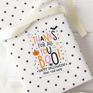 Pink, orange, and black Halloween gift tag displayed on a wrapped present, with candy corn, ghosts, a smiling pumpkin, bat, spider, and scattered stars.