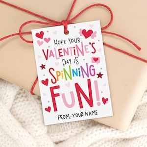 May include: A Valentine's Day gift tag with a red string bow. The tag reads "Hope Your Valentine's Day Is Spinning Fun!" in colorful, playful lettering, with heart and star accents. The tag includes space for a name.