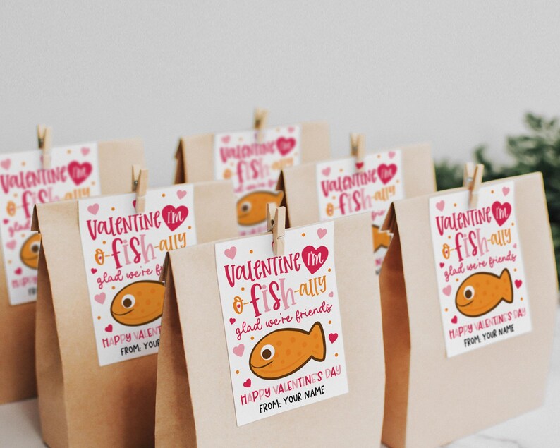 Kids classroom valentine printable tag that reads “You are o-fish-ally awesome” with “Happy Valentine’s Day” and “From: Your Name,” decorated with a smiling pink fish and scattered hearts, designed to be printed, cut, and attached to brown paper gift bags or snack favors.