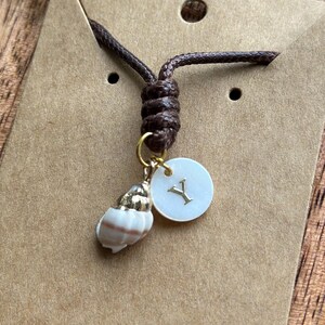 May include: A brown cord necklace featuring a small seashell pendant, a gold-colored charm, and a white circular charm with the letter "Y". The necklace is displayed on a brown card.