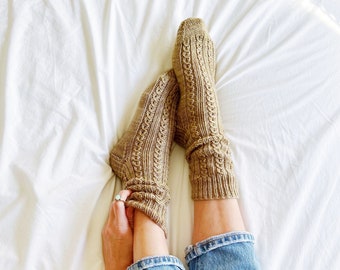 Può includere: Un paio di calzini di lana marroni con un motivo a treccia. I calzini sono indossati da una persona che indossa jeans blu.