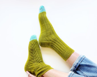 Peut inclure: Une paire de chaussettes en tricot vert avec des orteils mouchetés bleu et blanc. Les chaussettes sont portées par une personne portant un jean bleu et un bracelet avec des pierres vertes.