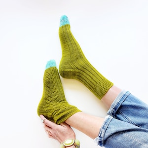 May include: A pair of green knitted socks with blue and white speckled toes. The socks are being worn by a person wearing blue jeans and a bracelet with green stones.