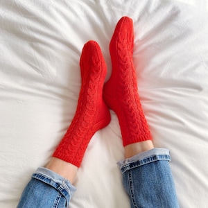 May include: A pair of red cable knit socks against a white background. The socks are being worn by someone in blue jeans.