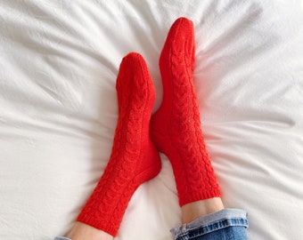May include: A pair of red cable knit socks with a white background. The socks are worn by a person wearing blue jeans.