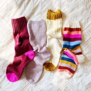 Könnte beinhalten: Vier Paar gestrickte Socken in verschiedenen Farben und Mustern. Die Socken liegen auf einer weißen, strukturierten Oberfläche. Die Socken umfassen ein Paar lila Socken, ein Paar weiße Socken mit einem goldenen Band, ein Paar rote Socken mit einem rosa Band und ein Paar mehrfarbige gestreifte Socken.