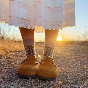 May include: A person wearing brown suede clogs with a buckle detail, paired with patterned socks. The socks are beige with a dark floral design. The person is wearing a white dress with a lace trim. The background is a field with the sun setting.