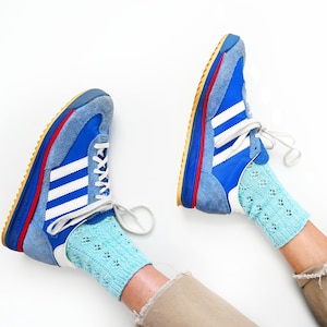 Peut inclure: Une paire de baskets bleu vif avec des bandes latérales blanches, des empiècements en daim bleu clair et des lacets blancs. Les baskets présentent une semelle intermédiaire rouge et bleue et sont portées avec des chaussettes en tricot bleu clair.
