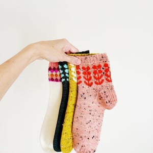 Puede incluir: Cuatro pares de calcetines de punto en color crema, negro, amarillo mostaza y rosa moteado. El par crema tiene un puño geométrico colorido, el negro un patrón azul, el amarillo un puño de espiga blanco y el rosa un motivo floral rojo.