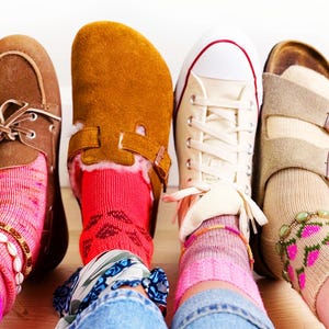 May include: Four pairs of feet wearing different footwear: a brown suede boat shoe with pink socks, a tan suede clog with red socks, a white canvas trainer with striped socks, and a tan double-strap sandal with patterned socks and an anklet.
