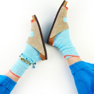 Może przedstawiać: Jasnobrązowe sandały zamszowe z korkową podeszwą, noszone z jasnoniebieskimi skarpetkami z pomarańczowym wykończeniem. Jeden kostek zdobi ozdobna bransoletka z niebieskimi i złotymi akcentami. Na białym tle.