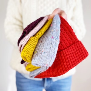 May include: A stack of four knitted hats in different colors: red, gray, yellow, and pink. The hats are folded and stacked on top of each other.
