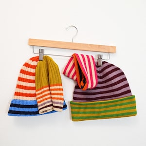 May include: Three striped knit hats hanging on a wooden hanger. The hats are in shades of blue, orange, yellow, green, and purple.