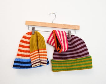 Puede incluir: Tres gorros de punto a rayas colgados de una percha de madera. Los gorros son en tonos de azul, naranja, amarillo, verde y morado.