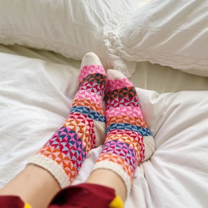 May include: A pair of colourful knitted socks with a geometric pattern in shades of pink, orange, blue, and white. The socks are being worn by someone sitting on a white bed.