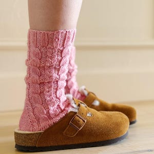 May include: A pair of brown suede clogs with a buckle closure, worn with pink cable-knit socks. The socks are a light pink color and have a textured, woven design. The clogs have a cork sole and a shearling lining.