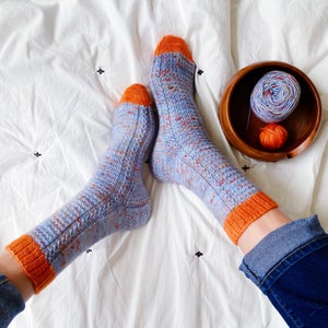 May include: A pair of blue and orange knitted socks with a cable pattern. The socks are on a white bedspread with a bowl of yarn in the background.