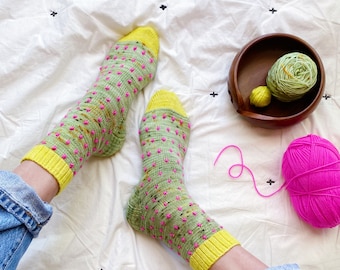 May include: A pair of green and yellow socks with pink polka dots. The socks are being worn by a person with jeans. There are balls of yarn in the background.