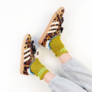 May include: A pair of leopard print trainers with white stripes and a brown sole. The trainers are worn with a pair of socks that are green with a thin blue stripe. The person is wearing grey sweatpants.