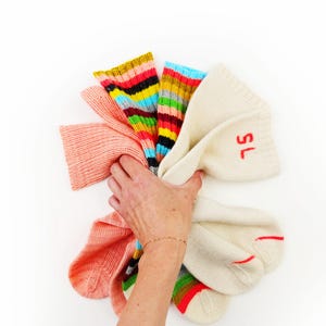 Puede incluir: Una mano sosteniendo una colección de calcetines coloridos. Los calcetines incluyen un par de calcetines rosa claro, un par con rayas arcoíris y un par de calcetines color crema con las letras "SL" en rojo. Los calcetines son de punto suave.
