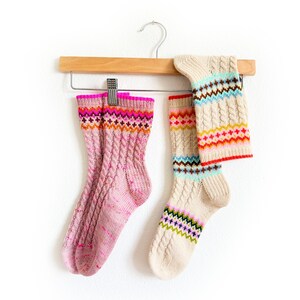 Könnte beinhalten: Zwei Paar bunte Stricksocken hängen an einem Holzbügel. Ein Paar ist rosa mit Zopfmuster und Bündchen. Das andere Paar ist cremefarben mit ähnlichem Zopfmuster und gemusterten Bündchen. Die Socken haben verschiedene Farben.