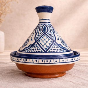 Op de afbeelding: Een keramische tajinepot met een conisch deksel en een brede basis. Het deksel is wit met blauwe geometrische patronen en een donkerblauwe knop. De basis is bruin en de rand is versierd met blauwe en witte accenten. Een traditioneel kookvat.