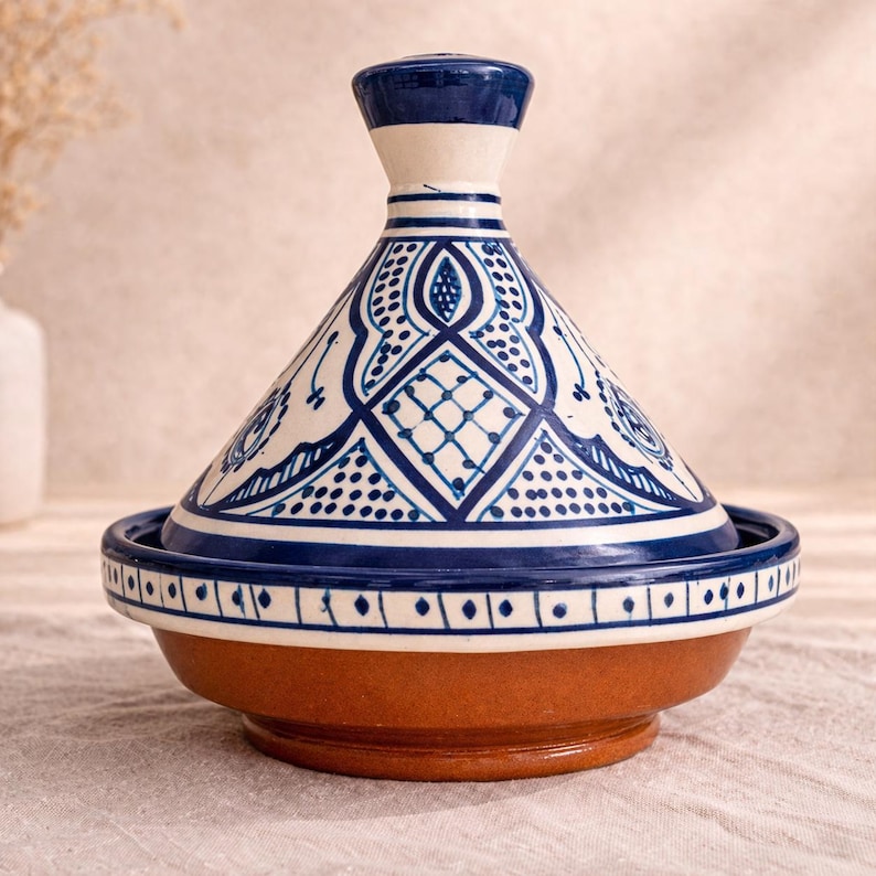 Op de afbeelding: Een keramische tajinepot met een conisch deksel en een brede basis. Het deksel is wit met blauwe geometrische patronen en een donkerblauwe knop. De basis is bruin en de rand is versierd met blauwe en witte accenten. Een traditioneel kookvat.