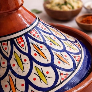 May include: A close-up of a traditional Moroccan tagine pot. The lid is decorated with a blue and white pattern, accented with orange, yellow, and red details. The pot is made of terracotta and is designed for slow cooking.