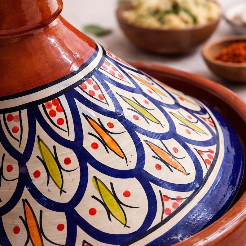 May include: A close-up of a traditional Moroccan tagine pot. The lid is decorated with a blue and white pattern, accented with orange, yellow, and red details. The pot is made of terracotta and is designed for slow cooking.