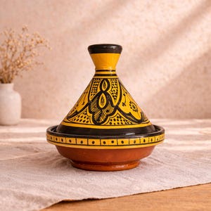May include: A ceramic tagine pot with a conical lid. The pot has a yellow and black design with intricate patterns. The base is reddish-brown, and the pot sits on a neutral-coloured surface.