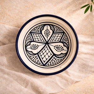 Op de afbeelding: Een ronde, witte keramische schaal met een donkerblauwe rand en ingewikkelde zwart-witte geometrische patronen in het midden. De schaal staat op een beige doek.