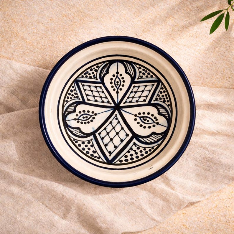 Op de afbeelding: Een ronde, witte keramische schaal met een donkerblauwe rand en ingewikkelde zwart-witte geometrische patronen in het midden. De schaal staat op een beige doek.