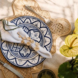 May include: A white ceramic plate with blue floral patterns, topped with a white napkin featuring a green stitched edge and a circular woven napkin ring. A silver fork and knife rest on a scalloped, light brown woven straw placemat.