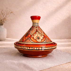 Puede incluir: Una olla de tagine de cerámica con tapa cónica y base ancha y poco profunda. La olla es principalmente marrón con patrones decorativos naranjas, rojos y crema. La tapa tiene un pomo rojo. El tagine está sobre un paño blanco.