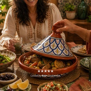 May include: A terracotta tagine with a decorative blue and white lid is being opened to reveal a meal. The tagine sits on a wooden table surrounded by bowls of food and drinks. The text "Loved by 600+ Customers" is visible.