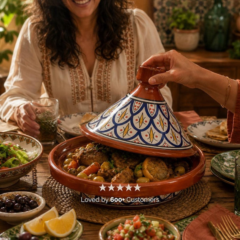 May include: A terracotta tagine with a decorative blue and white lid is being opened to reveal a meal. The tagine sits on a wooden table surrounded by bowls of food and drinks. The text "Loved by 600+ Customers" is visible.