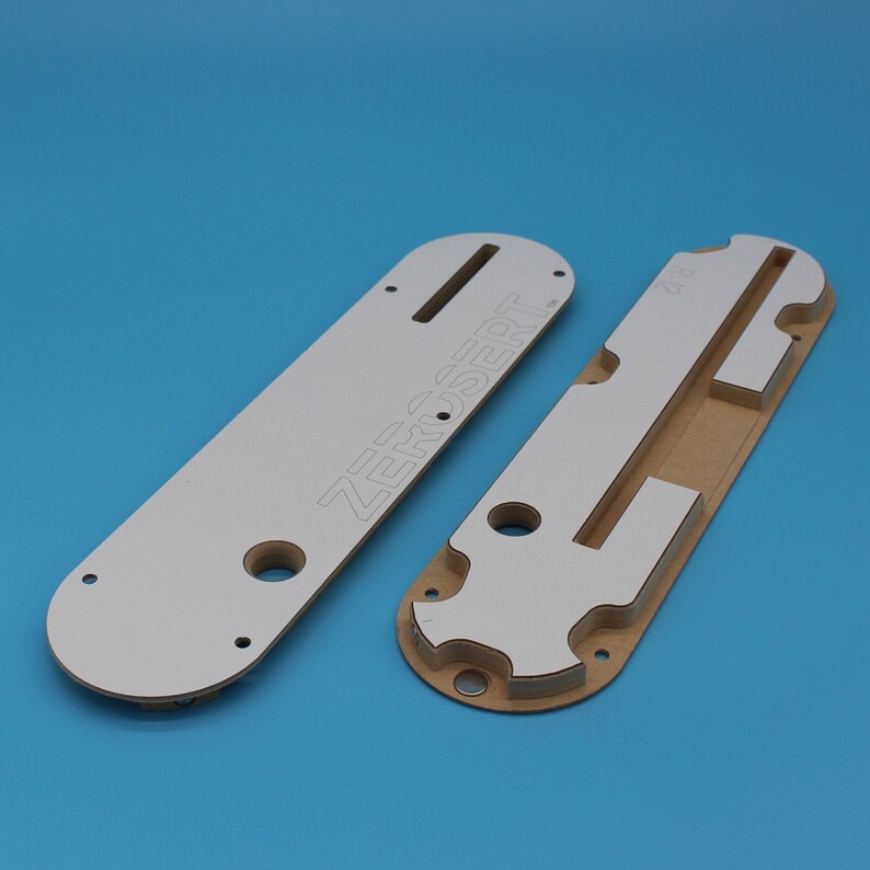 Puede incluir: Dos plantillas de fresado de madera blancas y marrones. La plantilla m&aacute;s grande tiene el texto "ZEROSERT" impreso en ella.