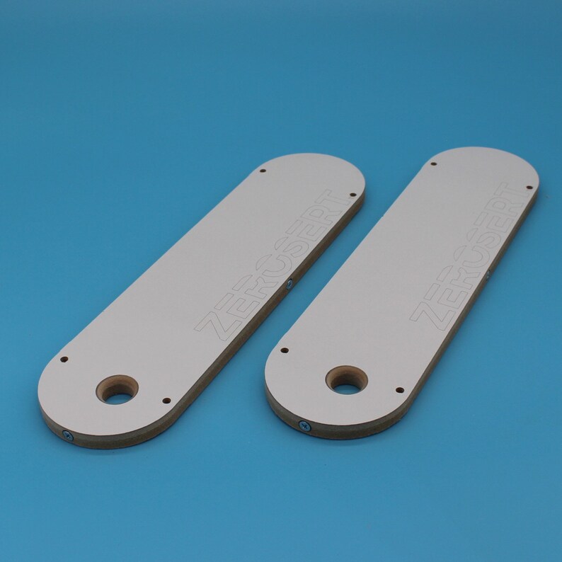 May include: Two white wooden router templates with the text "ZERO INSERT" printed on them. The templates have a large hole in the centre and several smaller holes around the edges.
