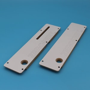 May include: Two white wooden router jigs with the text "ZERCSERT" printed on them. The jigs have multiple holes and a slot for a router bit.